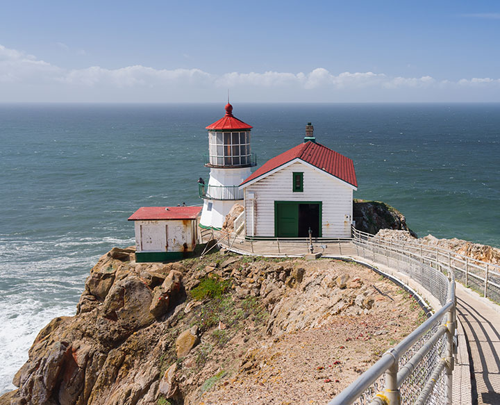 in the foreground, a trail with steel railings leads to a lighthouse on a cliff ledge, the lighthouse has a short light tower with a garage and small shed nearby, white walls, green trim and bright brick red shingles make up the structures